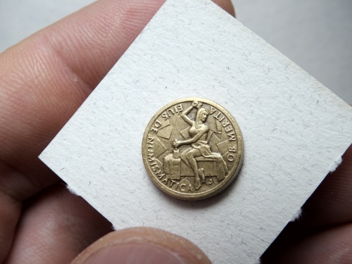  Klopový odznak s vyobrazením ženské postavy razící minci a opisem OB MERITA EIUS DE NUMISMATICA 